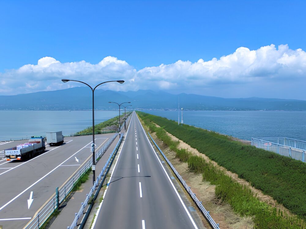 長崎県の諫早湾干拓堤防道路（雲仙多良シーライン）。青空の下、海を両側にまたいで真っ直ぐに伸びる見晴らしの良い直線道路。信号や交差点、対向車とのすれ違いがなく車線も広いため、ペーパードライバーが運転の基本感覚を取り戻すための最初の練習場所として最適。