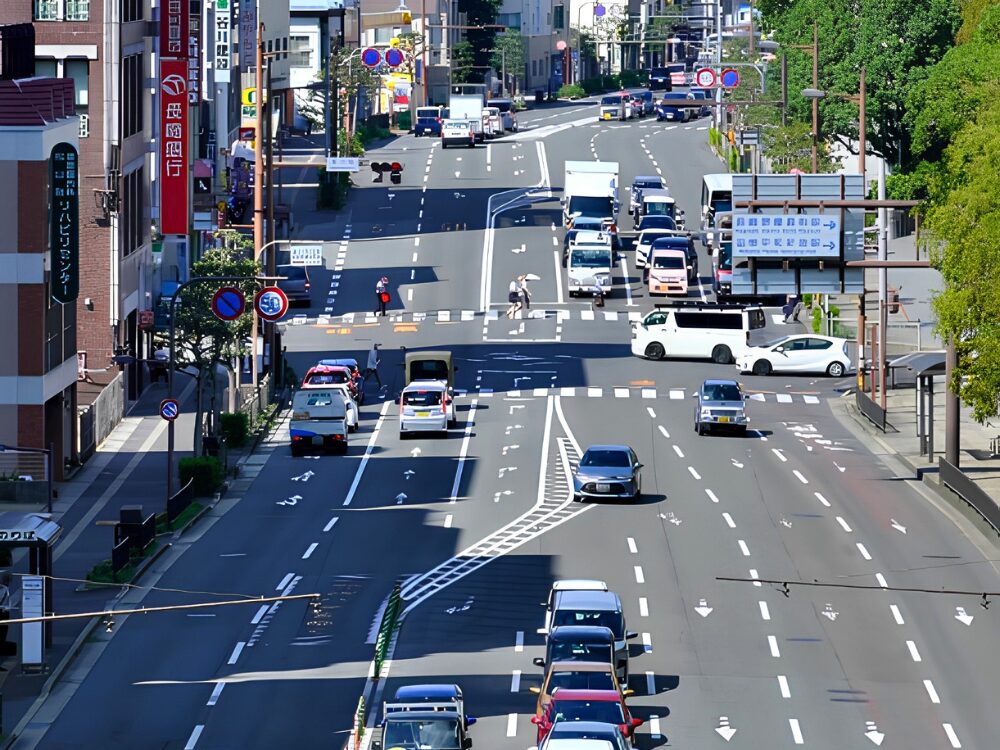 長崎特有の斜面市街地を背景に、緩やかに続く坂道の多車線道路を俯瞰した光景。多くの車がスムーズに行き交っており、日常的な交通状況がわかる。この風景に慣れることが、長崎での安心安全な運転への第一歩。